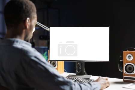 Téléchargez les photos : L'homme examine l'affichage blanc sur le poste de travail pendant le télétravail et utilise un écran vide sur l'écran. Homme entrepreneur opère à distance de la maison avec disposition chromatique vierge sur son bureau. - en image libre de droit