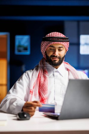 A focused Arabian businessman in Islamic attire sits at a modern desk, typing on a laptop. He efficiently works, utilizing wireless technology for online communication, surfing the net, and conducting