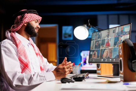 Foto de Empresario árabe confiado en la ropa tradicional dedicado a la videoconferencia en línea en un escritorio. Chico musulmán escucha atentamente a sus colegas, el uso de PC de escritorio para la comunicación y la investigación. - Imagen libre de derechos