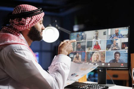 Serious Arab businessman holding paperwork and discussing ideas with his team through a video call on the desktop pc. Muslim guy comparing information with coworkers in an online meeting.