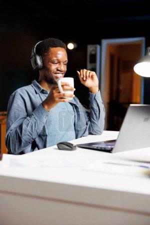 Foto de Chico afroamericano resolviendo tareas de economía en línea, divirtiéndose escuchando podcast en auriculares en casa. Freelancer masculino que trabaja en estrategia de gestión global con inversiones en fondos de capital. - Imagen libre de derechos