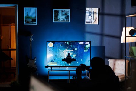 Photo for Player participates in online roleplaying firing match, hooking console through TV and enjoying nighttime gaming activity with friends. Male student is playing shooter video games. - Royalty Free Image