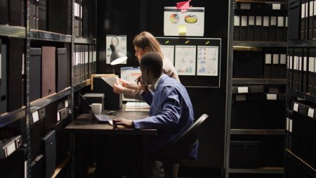 Interracial police partners work as private detectives, reviewing evidence in archive room for clues to a crime. Serious caucasian female inspector works with focused male investigator on case files.
