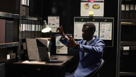 Black policeman investigates complex case, surrounded by evidence, files, and holographic displays. Private detective using artificial intelligence to examine evidence on hologram in incident room.