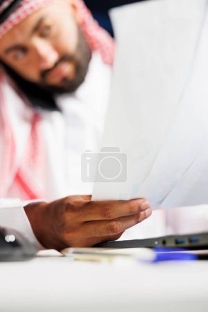 Enfoque selectivo en los documentos en poder de un joven vestido con ropa árabe y con una llamada telefónica. Primer plano de un musulmán centrado en examinar su papeleo mientras habla en un dispositivo móvil.