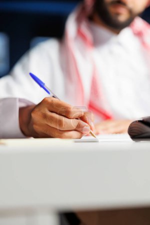 Photo for A focused businessman of Middle Eastern ethnicity efficiently works at his home office desk, taking research notes using a blue pen. Selective focus an Arab person writing down on his notebook. - Royalty Free Image