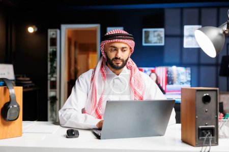 Arab guy focused on his laptop efficiently surfing the internet. Young man wearing Islamic clothing embodies productivity in the digital era, using his personal computer on the table.