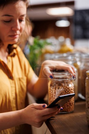 Foto de Mujer vegana en una dieta utilizando el teléfono para asegurarse de que los alimentos de supermercado cero residuos es bueno para su salud. Cliente en la tienda de comestibles del vecindario local asegurándose de que los productos sean naturales - Imagen libre de derechos