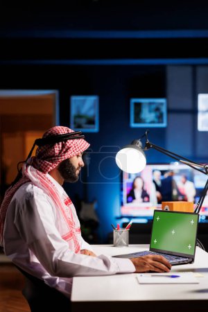 Photo for Arab male professional wearing traditional attire is seated at an office desk, using a laptop with an empty green screen. The image portrays a modern workspace for advertising or presentations. - Royalty Free Image