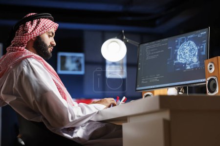 Foto de El hombre islámico utiliza la IA y el aprendizaje automático para garantizar la seguridad cibernética de una red. Supervisa los centros de datos y servidores que trabajan con un monitor de PC que muestra software avanzado y una red neuronal. - Imagen libre de derechos