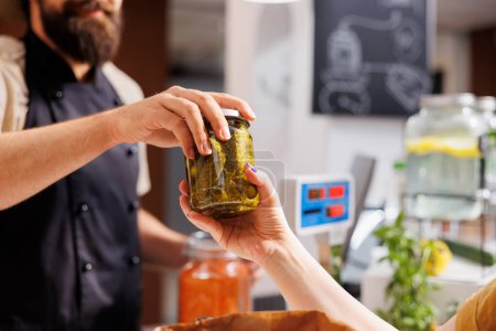 Photo for Close up shot of customer at checkout counter in zero waste shop, buying locally sourced ecofriendly vegetables from storekeeper. Client purchasing pickles jar in local neighborhood store - Royalty Free Image