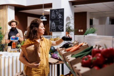Woman in zero waste shop purchasing organic locally grown vegetables, picking ripe eggplants. Customer in local grocery shop looking to buy healthy food, using paper bag to avoid single use plastics