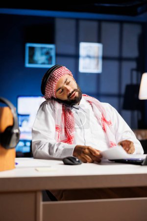Empresario musulmán con atuendo tradicional, hablando en su teléfono celular y revisando notas en un portátil. Empresario árabe conversando con un compañero de trabajo y comparando la investigación sobre un minicomputador.