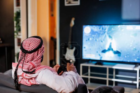 Arab man is concentrated and immersed, he controls the gameplay using a console controller. The digital world comes alive on his television display as he grasps wireless technology.