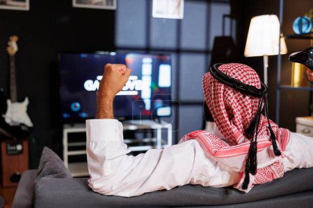 Detailliertes Bild eines arabischen Mannes, der seinen Sieg mit der Faust in der Luft feiert, während er auf einer bequemen Couch sitzt. Rückansicht eines Mannes aus dem Nahen Osten, der einen drahtlosen Controller greift.
