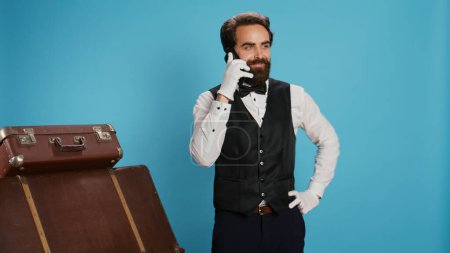 Doorman talks on phone call with hotel guests, making room reservations against blue background. Professional bellhop chatting with visitors on camera and accepting remote bookings.