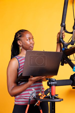 Joven dama negra usando las pautas de dispositivo electrónico para examinar cuidadosamente y arreglar su bicicleta rota. Mujer deportiva utiliza ordenador portátil y herramientas especializadas para examinar modificar y reparar la bicicleta.