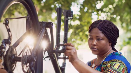 Determined black woman testing and inspecting bike wheel and pedals in yard for summer cycling. African american female cyclist with caucasian man outdoor repairing bicycle parts using work tools.