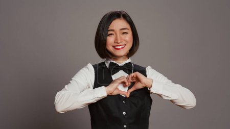 Photo for Hotel administrator shows heart shape symbol on camera, expressing romantic gesture and being flirty. Asian receptionist presenting true honest feelings, does romance sign in studio. Camera B. - Royalty Free Image