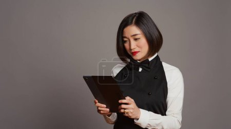 Photo for Asian restaurant server checking all table reservations on tablet, browsing through clients information on website. Waitress looking at online webpages to see availability. Camera B. - Royalty Free Image