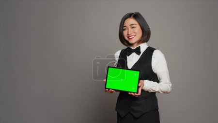 Téléchargez les photos : Asiatique réceptionniste tenant tablette avec écran vert sur la caméra, présentant modèle de maquette sur gadget moderne. Concierge de l'hôtel regardant l'affichage de la mise en page de copyspace, studio gris. Caméra A. - en image libre de droit