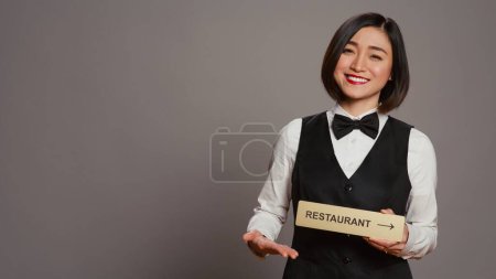 Téléchargez les photos : Concierge hôtel asiatique tenant signe restaurant pour indiquer la direction, pointant vers la salle à manger. Réceptionniste aidant les clients à profiter de toutes les commodités, se tient sur fond gris. Caméra B. - en image libre de droit