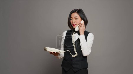 Photo for Asian receptionist answering landline phone call in studio, talking on vintage telephone with cable. Woman front desk staff using phone with cord to confirm room reservation. Camera A. - Royalty Free Image