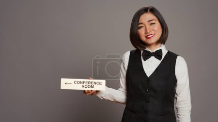 Photo for Asian receptionist pointing at wall indicator for conference room, using pointer to help customers find all amenities and facilities. Woman employee holding sign to indicate directions. Camera B. - Royalty Free Image