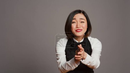 Photo for Restaurant hostess clapping hands and celebrating for someone, cheering and saying congratulations in studio. Asian waitress with uniform and apron applauding a person, acclaim. Camera B. - Royalty Free Image