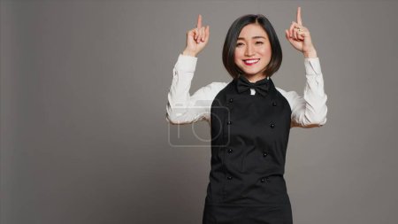 Photo for Asian woman pointing at something upwards in studio, indicating direction above her head. Waitress server with apron showing up indication over grey background, table service. Camera A. - Royalty Free Image