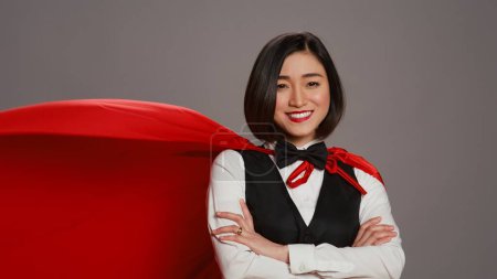 Photo for Hotel administrator wearing a red hero cloak on camera, feeling strong and powerful to help clients. Asian receptionist posing with confidence using a superhero cape over grey background. Camera B. - Royalty Free Image