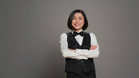 Téléchargez les photos : Femme asiatique concierge d'hôtel portant un élégant uniforme avec arc, travaillant dans l'industrie hôtelière en tant que réceptionniste. Administrateur de réception saluant les clients, fond gris. Caméra A. - en image libre de droit