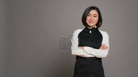Restaurant hostess posing with confidence in studio, standing with arms crossed over grey background. Asian waitress barista with gourmet serving expertise smiling on camera. Camera A.