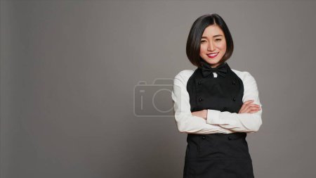 Photo for Asian diner employee prepared to serve food at tables, working in hospitality cuisine and catering. Restaurant waitress with apron standing over grey background, gourmet service. Camera A. - Royalty Free Image