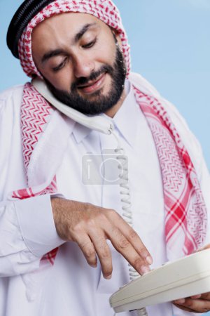 Foto de Hombre musulmán con pañuelo tradicional marcando el número en el teléfono fijo. Árabe vestido con ropa cultural islámica sosteniendo teléfono retro y haciendo una llamada de primer plano - Imagen libre de derechos