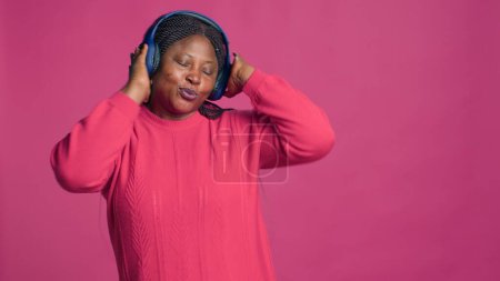 Modebloggerin mit kabellosen Kopfhörern und Spaß am Rhythmus der Lieblingsmusik. Atemberaubende schwarze Frau im rosa Pullover tanzt unbeschwert und genießt die angenehme Melodie.