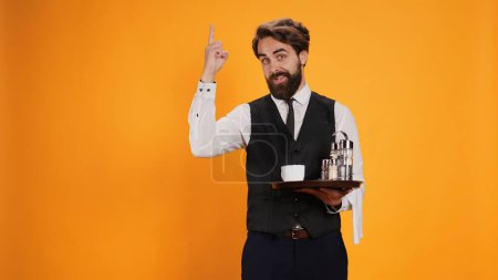 Photo for Qualified worker in studio having new idea, standing with food tray in hand and raising index finger upwards. Competent butler carrying cutlery and drink to serve people, restaurant sector. - Royalty Free Image