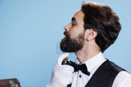 Photo for Hotel worker sprays cologne to smell fresh, applying perfume in studio and feeling excellent while he poses in formal attire with gloves. Doorman spraying aromatic scent. Close up. - Royalty Free Image