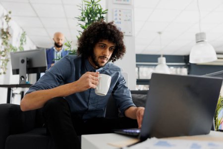 Young arab comapny employee drinking coffee and using corporate laptop in business office. Executive manager holding tea mug and solving work tasks on computer in coworking space
