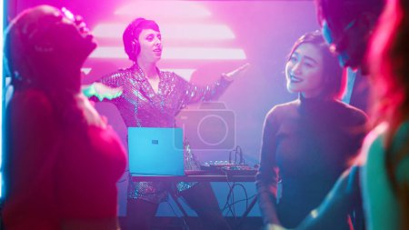 Téléchargez les photos : Funky DJ féminin mixant des sons sur scène au club, dansant avec un groupe d'amis à la station audio. Jeune femme sautant avec des gens divers, profiter de la nuit à la soirée de boîte de nuit. Trépied. - en image libre de droit
