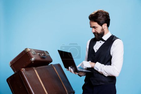 Amistoso portero con computadora portátil en la mano, la publicidad de ocupación profesional en la industria hotelera. Decidido botones feliz con corbata y guantes, presentando la hospitalidad en la cámara.