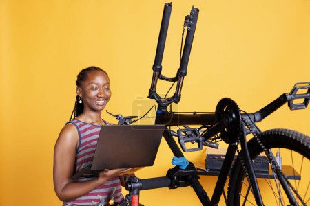 Photo for Female cyclist of african american ethnicity fixes damaged bike using toolkit and laptop to research solutions. Using minicomputer and professional equipment, black woman is repairing broken bicycle. - Royalty Free Image