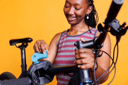 Foto de Vista detallada de la mujer negra examinando el marco dañado de la bicicleta en el soporte de reparación contra el fondo amarillo. Mujer afroamericana asegurando y haciendo los ajustes necesarios en la bicicleta moderna. - Imagen libre de derechos