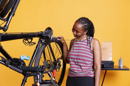 Dedicated black woman checking bicycle tires for damages to fix with professional work tools. Active african american female working on broken bike rubber wheel with specialized equipment.