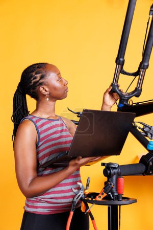 Young black lady does bike maintenance in front of yellow background, using digital laptop for instructions. African american woman repairing bicycle with personal computer and specialized tools.