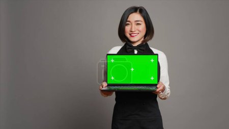 Photo for Asian server presenting greenscreen display on laptop in studio, wearing uniform with apron and bow. Restaurant hostess holding pc with blank mockup template, copyspace layout. Camera A. - Royalty Free Image