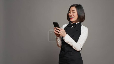 Photo for Restaurant hostess texting messages to confirm reservations on phone app, working in catering and serving industry. Asian waitress using smartphone online websites, social media. Camera A. - Royalty Free Image