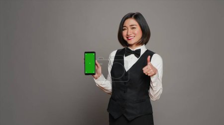 Foto de Conserje de hotel asiático apuntando al teléfono con diseño de pantalla verde, sosteniendo el dispositivo que ejecuta la plantilla de cromakey aislado. Recepcionista que muestra la pantalla del teléfono inteligente con la maqueta de copyspace. Cámara A. - Imagen libre de derechos