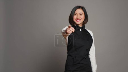 Photo for Asian waitress pointing at camera to choose you over grey background, wearing uniform with bow and apron. Woman catering worker choosing someone to work at a restaurant. Camera A. - Royalty Free Image
