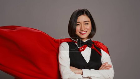 Téléchargez les photos : La réceptionniste asiatique posant avec une cape de super-héros rouge à la caméra, se sentant confiante et puissante alors qu'elle porte un manteau en héros. Concierge de l'hôtel debout les bras croisés. Caméra B. - en image libre de droit
